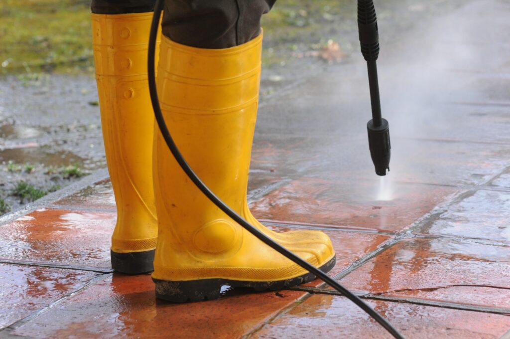 A person wearing yellow rubber boots with high-pressure water nozzle cleaning the dirt in the tiles