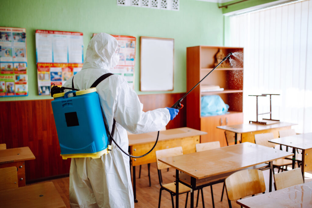 Back view of a professional sanitary worker disinfecting the classroom before the academic year starts. A man wearing protective suit cleans up the auditorium from coronavirus Covid-19. Health care